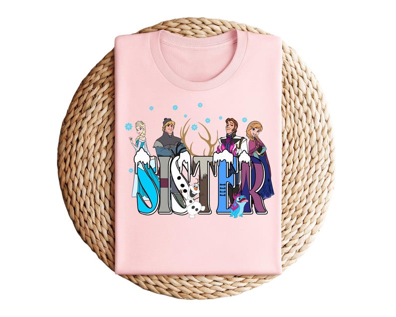 May include: A light pink t-shirt featuring the word "SISTER" in a colorful, winter-themed design. The design includes cartoon characters and snowflakes. The shirt is folded on a woven, circular surface.