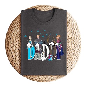 May include: A dark gray t-shirt featuring the word "DADDY" in a stylized font with characters from the movie Frozen. The letters are decorated with images of Elsa, Anna, Kristoff, Olaf, and Sven. Blue snowflakes are scattered above the letters.