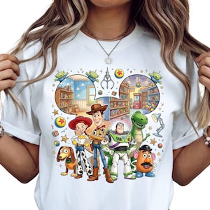 Pode incluir: Camiseta branca com um gr&aacute;fico colorido dos personagens de Toy Story, incluindo Woody, Buzz Lightyear e Rex. O design inclui uma m&aacute;quina de garra e outros elementos tem&aacute;ticos. A camiseta &eacute; de gola redonda.