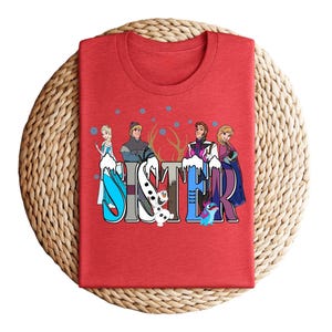 May include: Red t-shirt featuring the word "SISTER" in a colorful design with cartoon characters. The design includes characters from the movie Frozen, such as Elsa, Anna, Kristoff, and Olaf. The shirt is folded on a woven surface.