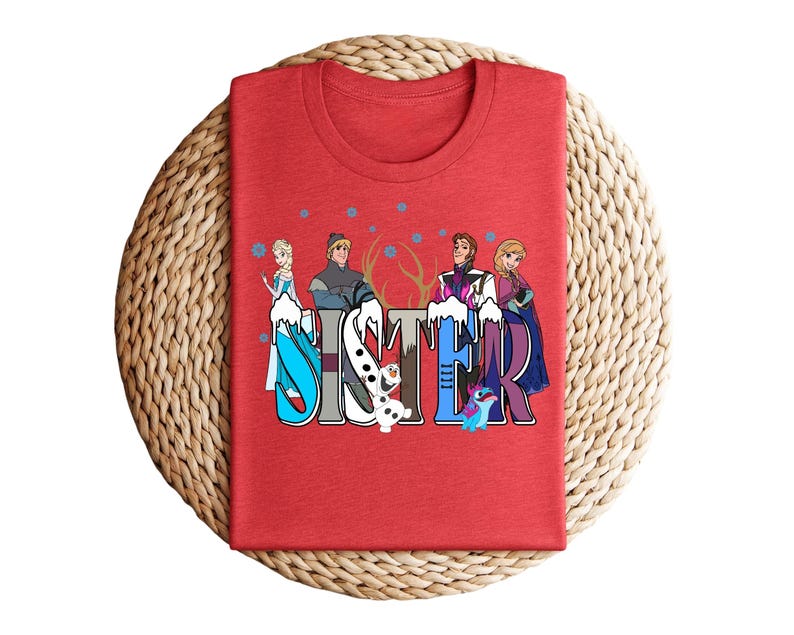 May include: Red t-shirt featuring the word "SISTER" in a colorful design with cartoon characters. The design includes characters from the movie Frozen, such as Elsa, Anna, Kristoff, and Olaf. The shirt is folded on a woven surface.