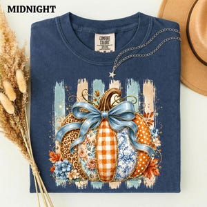 May include: A dark blue t-shirt featuring a graphic of a pumpkin decorated with leopard print, orange gingham, and blue floral patterns. A large light blue bow sits atop the pumpkin, which is surrounded by autumn leaves and blue flowers.