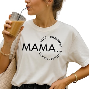 May include: White t-shirt with the word "MAMA" in large black letters. The word is surrounded by a circle of words including "LOVED", "HARDWORKING", "SELFLESS", and "PROTECTIVE". The person is holding a clear plastic cup with a straw.