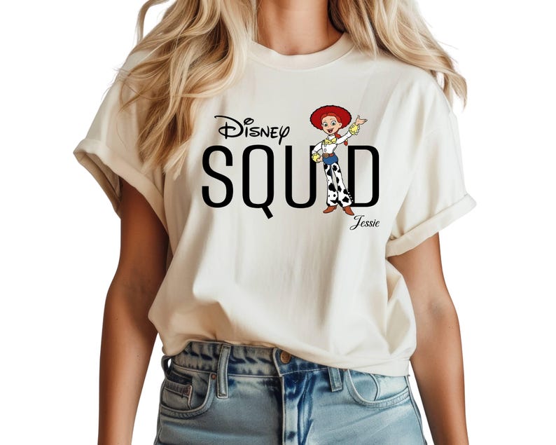 Puede incluir: Camiseta color crema con la frase "Disney SQUAD" en negro y una imagen de dibujos animados de Jessie de Toy Story. La palabra "Jessie" est&aacute; impresa debajo del personaje. La camiseta tiene las mangas enrolladas.