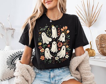 May include: A black short-sleeve t-shirt featuring a graphic print of five white ghosts, one wearing a gold crown, surrounded by colorful autumn flowers, green leaves, and a small bat. The design is centered on the chest of the garment.