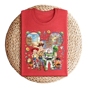 Pode incluir: Camiseta vermelha com um gr&aacute;fico colorido de personagens de Toy Story, incluindo Woody, Buzz Lightyear, Jessie, Rex, Sr. Batata e Slinky. O design incorpora cenas do filme. A camiseta est&aacute; dobrada sobre uma superf&iacute;cie tecida.