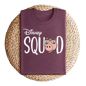 Puede incluir: Una camiseta granate con el texto "Disney SQUAD" en blanco. La "O" de SQUAD se reemplaza con un cerdo de dibujos animados con gafas de sol. La camiseta est&aacute; doblada sobre un salvamanteles circular tejido.
