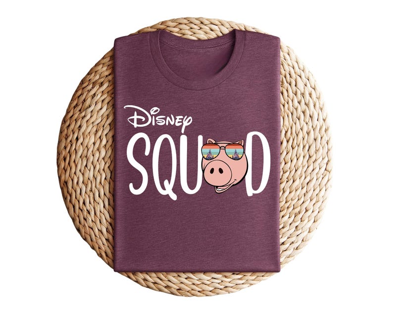 Puede incluir: Una camiseta granate con el texto "Disney SQUAD" en blanco. La "O" de SQUAD se reemplaza con un cerdo de dibujos animados con gafas de sol. La camiseta est&aacute; doblada sobre un salvamanteles circular tejido.