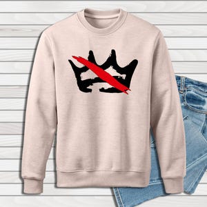 Peut inclure: Un sweat-shirt beige clair &agrave; col rond avec un motif de couronne noire barr&eacute;e d'une barre rouge. Le sweat-shirt est pr&eacute;sent&eacute; avec un jean en denim bleu. Le motif de la couronne est centr&eacute; sur le devant.