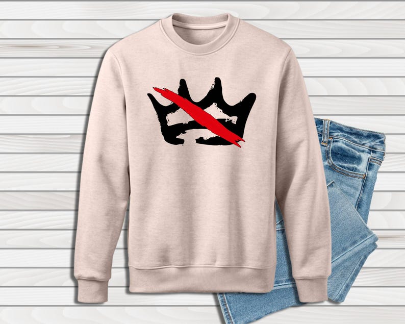 Peut inclure: Un sweat-shirt beige clair &agrave; col rond avec un motif de couronne noire barr&eacute;e d'une barre rouge. Le sweat-shirt est pr&eacute;sent&eacute; avec un jean en denim bleu. Le motif de la couronne est centr&eacute; sur le devant.