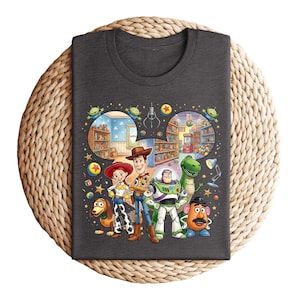 Pode incluir: Camiseta cinza escuro com um gr&aacute;fico colorido dos personagens de Toy Story, incluindo Woody, Buzz Lightyear e Rex. O design inclui cenas do filme, como a loja de brinquedos e a m&aacute;quina de garras. A camiseta &eacute; exibida em uma superf&iacute;cie circular tecida.