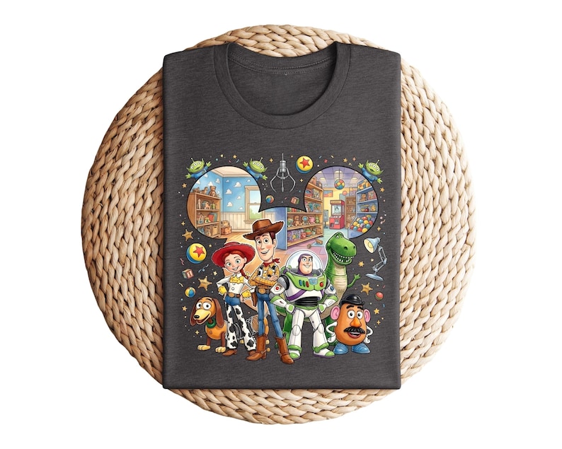 Pode incluir: Camiseta cinza escuro com um gr&aacute;fico colorido dos personagens de Toy Story, incluindo Woody, Buzz Lightyear e Rex. O design inclui cenas do filme, como a loja de brinquedos e a m&aacute;quina de garras. A camiseta &eacute; exibida em uma superf&iacute;cie circular tecida.