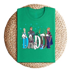 May include: A green t-shirt featuring the word "DADDY" in a stylized font with characters from the movie Frozen. The design includes Elsa, Kristoff, Anna, Olaf, and Sven. The shirt is displayed on a woven, circular surface.