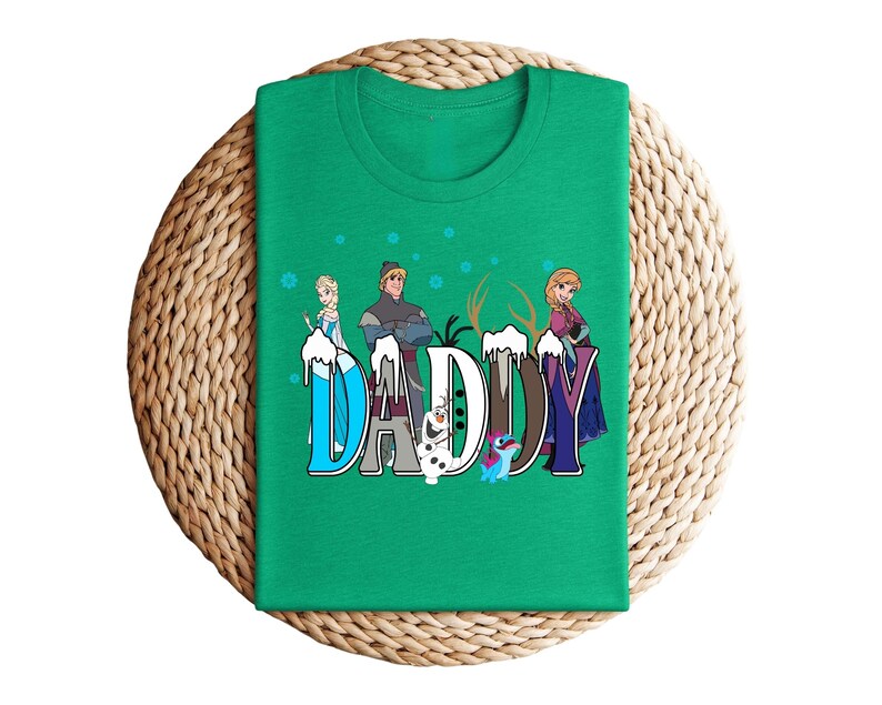 May include: A green t-shirt featuring the word "DADDY" in a stylized font with characters from the movie Frozen. The design includes Elsa, Kristoff, Anna, Olaf, and Sven. The shirt is displayed on a woven, circular surface.