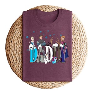 May include: A maroon t-shirt featuring the word "DADDY" in a stylized font with characters from the movie Frozen. The letters are decorated with snowflakes and images of Elsa, Anna, Kristoff, Olaf, and Sven.