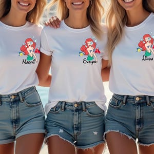 May include: Three white t-shirts with a cartoon mermaid design and names printed below. The mermaid has red hair, a green top, and is surrounded by bubbles and fish. The names printed are Naomi, Evelyn, and Linda. The shirts are paired with denim shorts.