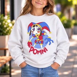 May include: A white long-sleeve sweatshirt featuring a graphic print of a cartoon jester character with large eyes, wearing a red and blue jester hat and outfit. The text POMNI is printed in red capital letters below the character illustration.