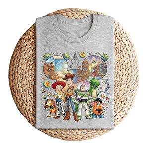 Pode incluir: Uma camiseta cinza claro com um gr&aacute;fico colorido de personagens da franquia de filmes Toy Story. O design inclui Woody, Buzz Lightyear, Jessie, Rex, Sr. Batata e Slinky Dog, junto com cenas de lojas de brinquedos.