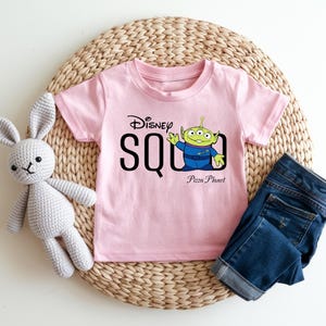 Puede incluir: Una camiseta rosa claro con el texto "Disney SQUAD" y un personaje de dibujos animados alien&iacute;gena de la pel&iacute;cula Toy Story. La camiseta tambi&eacute;n tiene las palabras "Pizza Planet" impresas debajo del alien&iacute;gena. Unos vaqueros azules y un conejito de peluche gris tambi&eacute;n est&aacute;n en la imagen.