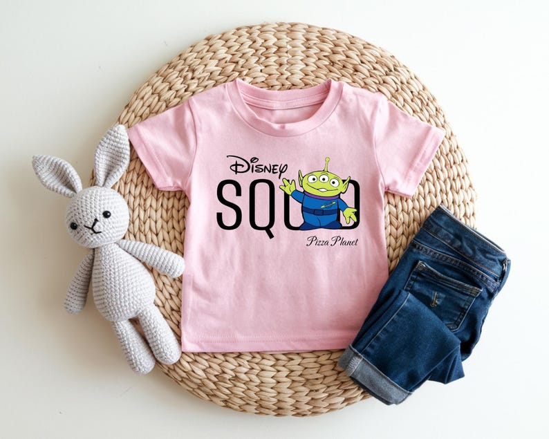 Puede incluir: Una camiseta rosa claro con el texto "Disney SQUAD" y un personaje de dibujos animados alien&iacute;gena de la pel&iacute;cula Toy Story. La camiseta tambi&eacute;n tiene las palabras "Pizza Planet" impresas debajo del alien&iacute;gena. Unos vaqueros azules y un conejito de peluche gris tambi&eacute;n est&aacute;n en la imagen.