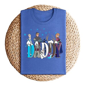 May include: A blue t-shirt featuring the word "DADDY" in a stylized font with characters from the movie Frozen. The design includes images of Elsa, Anna, Kristoff, and Olaf, with snowflakes and ice accents.