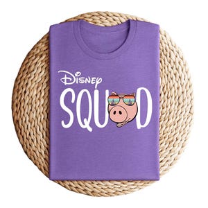 Puede incluir: Una camiseta morada con la frase "Disney Squad" en blanco. La "O" de "Squad" se reemplaza con un cerdo de dibujos animados con gafas de sol. La camiseta est&aacute; doblada y colocada sobre una estera circular tejida.