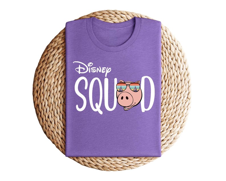 Puede incluir: Una camiseta morada con la frase "Disney Squad" en blanco. La "O" de "Squad" se reemplaza con un cerdo de dibujos animados con gafas de sol. La camiseta est&aacute; doblada y colocada sobre una estera circular tejida.