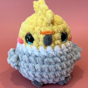 May include: A handmade, plush cockatiel bird in yellow, gray, and white. The bird has large, shiny black eyes with blue highlights, a small orange beak, and pink cheeks. The bird is made of soft, textured yarn.