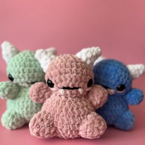 May include: Three handmade crochet monster toys in pastel colors. The toys are in shades of green, pink, and blue, with white horns, black eyes, and small fangs. The toys are sitting on a pink background.