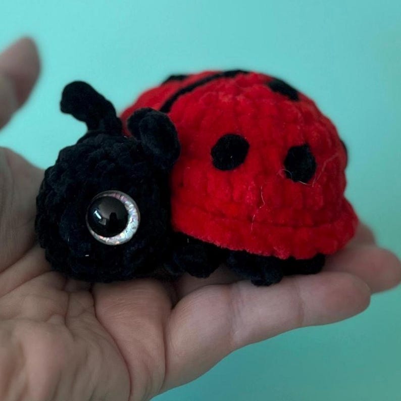 May include: A handmade plush ladybug toy. The ladybug has a red body with black spots and a black head. The toy is made of soft, textured material and has large, reflective eyes. The ladybug is a cute and whimsical item.