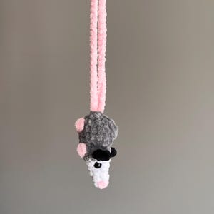 May include: A small, crocheted plush opossum hanging from a long, light pink yarn loop. The opossum features a fuzzy gray body, a white face with black eyes, small pink ears, and a pink nose. The texture is soft and yarn-based throughout.