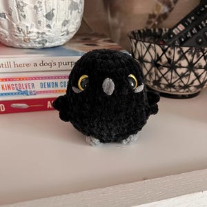 May include: A handmade black crochet owl with gold-colored eyes and gray accents. The owl has small wings and feet, and is sitting on a white surface. The owl is a decorative item.