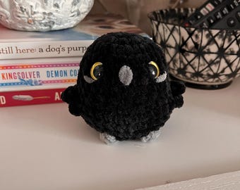 May include: A handmade black crochet owl with gold-colored eyes and gray accents. The owl has small wings and feet, and is sitting on a white surface. The owl is a decorative item.