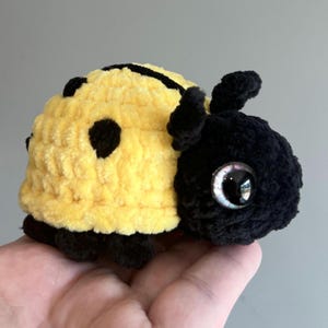 May include: A handmade crochet ladybug plushie. The plushie is yellow with black spots and a black head. It has large, shiny, iridescent eyes and small antennae. The ladybug is made of soft, textured yarn.