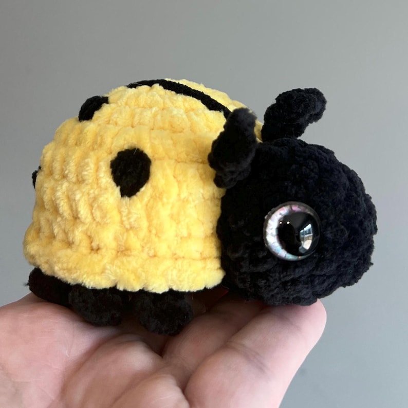 May include: A handmade crochet ladybug plushie. The plushie is yellow with black spots and a black head. It has large, shiny, iridescent eyes and small antennae. The ladybug is made of soft, textured yarn.
