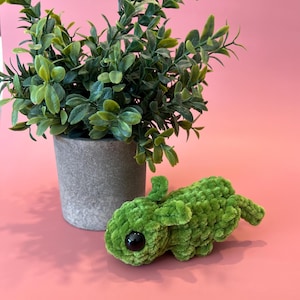 May include: A handmade green crochet bunny with black eyes rests on a pink surface next to a small potted plant with green leaves. The bunny is made of soft, textured yarn and has small ears and a tail.
