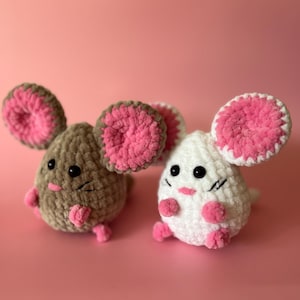 May include: Two crocheted mouse toys. One is brown, the other is white. Both have large pink and brown ears, small pink feet, and black eyes. The background is a solid pink.