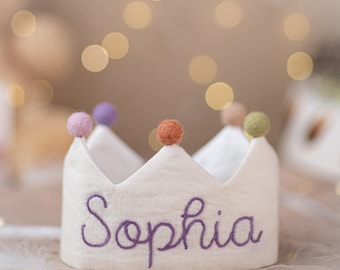 May include: A cream-colored fabric crown featuring the name Sophia embroidered in purple cursive thread. The crown is topped with five small, round, felted wool balls in various muted colors including pink, purple, orange, tan, and olive green.