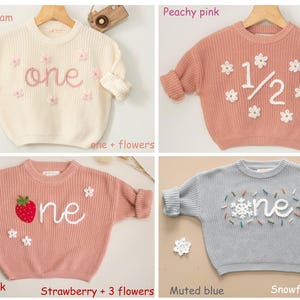 May include: Four different knit sweaters for babies, each with a different design and color. The sweaters are white with pink embroidery, peach with white embroidery, peach with red embroidery, and blue with white embroidery. The sweaters all say "one" on them.