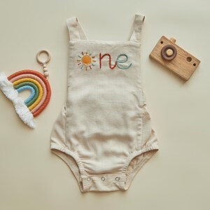 May include: A beige linen baby romper with embroidered sun and "one" text. A wooden toy camera and a rainbow-shaped toy with a white cloud accent are also present.