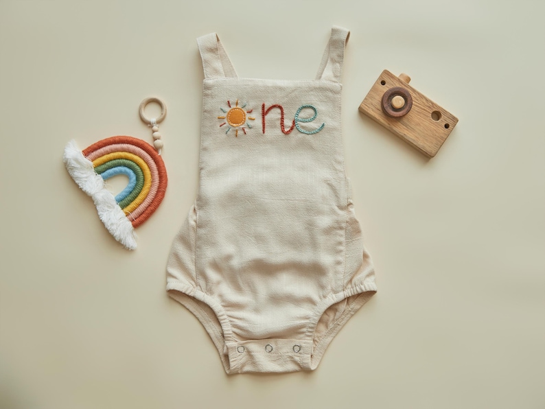 May include: A beige linen baby romper with embroidered sun and "one" text. A wooden toy camera and a rainbow-shaped toy with a white cloud accent are also present.