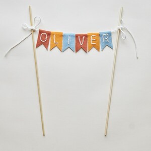 May include: A colorful cake topper with the name "OLIVER" spelled out on a banner. The banner is made of small, colorful flags in shades of red, orange, and blue. The flags are strung between two wooden dowels.