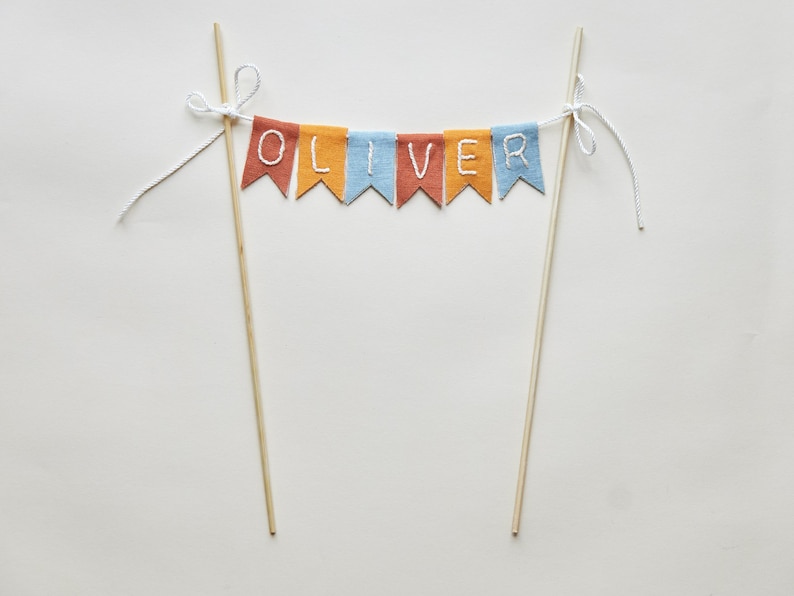 May include: A colorful cake topper with the name "OLIVER" spelled out on a banner. The banner is made of small, colorful flags in shades of red, orange, and blue. The flags are strung between two wooden dowels.