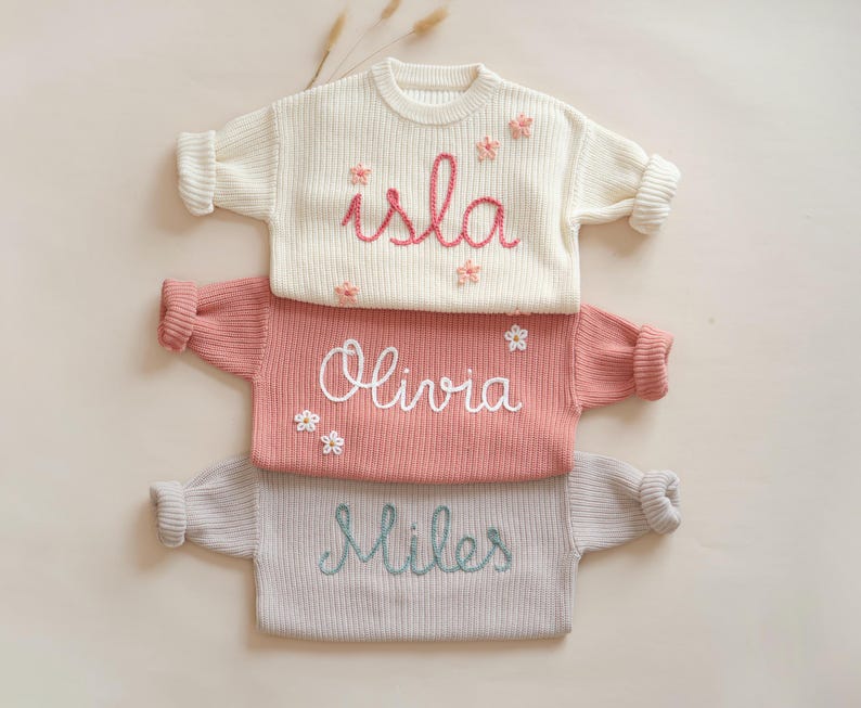 May include: Three knitted baby sweaters with embroidered names: Isla, Olivia, and Miles. Each sweater features a different color and has small embroidered flowers.