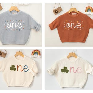 May include: Four different colored knitted baby sweaters with the word "one" embroidered on the front. The sweaters are in muted blue, clay terracotta, and two shades of white.