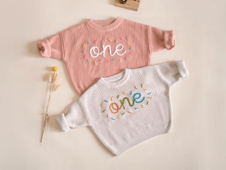May include: Two knitted baby sweaters with the word "one" embroidered on them. One sweater is pink and the other is white. The word "one" is embroidered in different colors on each sweater.