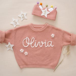 May include: A pink knitted jumper with the name "Olivia" embroidered on the front. The jumper has white embroidered flowers and stars on it. A pink crown with a white star and colourful pom-poms is also in the image.