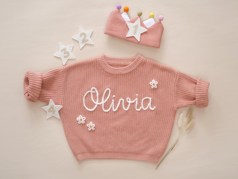 May include: A pink knitted sweater with the name "Olivia" embroidered on the front. The sweater has white embroidered flowers and stars on it. A pink crown with a white star and colorful pom-poms is also in the image.