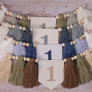 May include: A white felt banner with the number "1" embroidered on it four times. The banner is strung with wooden beads and brown, white, blue, and green tassels.