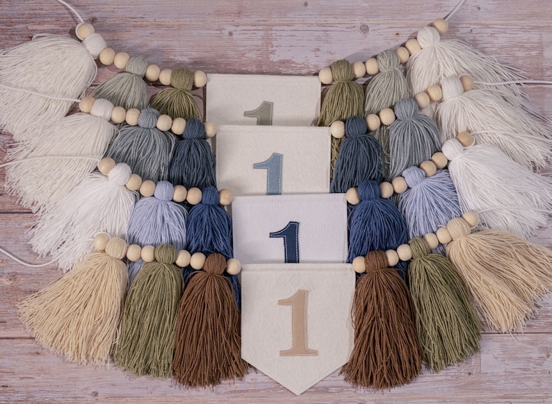May include: A white felt banner with the number "1" embroidered on it four times. The banner is strung with wooden beads and brown, white, blue, and green tassels.